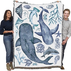 May include: A woven throw blanket with a marine life design. The blanket features sharks, fish, and jellyfish in blue and teal hues against a white backdrop. The blanket has a fringed edge. The blanket's dimensions are in centimetres.
