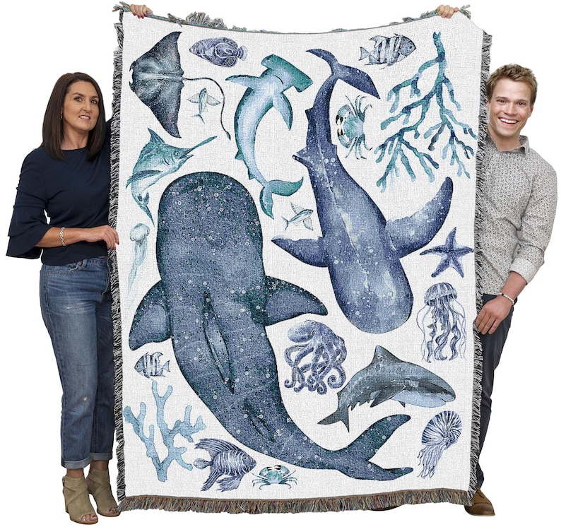 May include: A woven throw blanket with a marine life design. The blanket features sharks, fish, and jellyfish in blue and teal hues against a white backdrop. The blanket has a fringed edge. The blanket's dimensions are in centimetres.