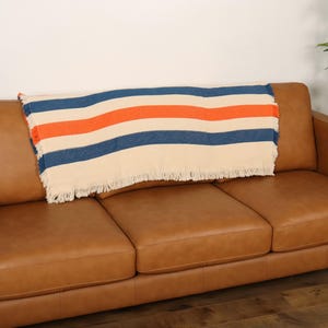 May include: A tan leather sofa with a striped blanket. The blanket has cream, navy blue, and orange stripes and a fringed edge. The sofa has three cushions and wooden legs.
