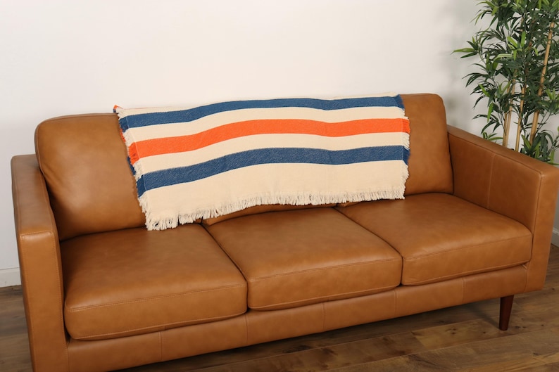 May include: A tan leather sofa with a striped blanket. The blanket has cream, navy blue, and orange stripes and a fringed edge. The sofa has three cushions and wooden legs.
