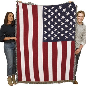 May include: A woven blanket featuring the American flag design. The blanket displays red and white stripes with a blue square in the upper left corner filled with white stars. The blanket has a fringed edge.
