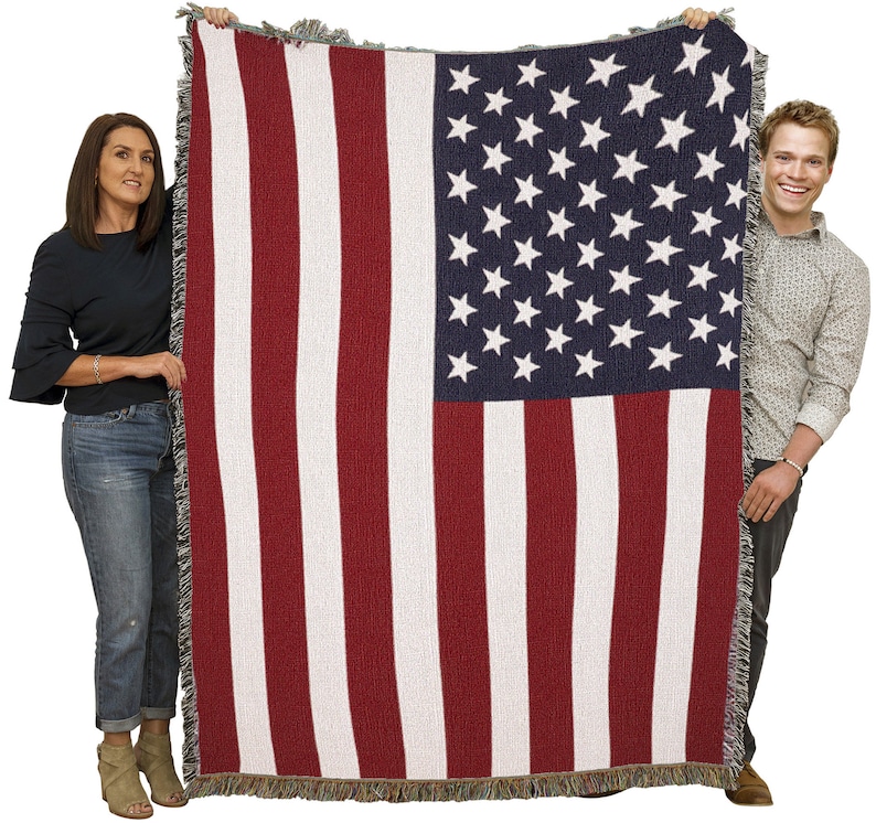 May include: A woven blanket featuring the American flag design. The blanket displays red and white stripes with a blue square in the upper left corner filled with white stars. The blanket has a fringed edge.
