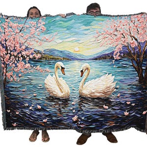 May include: A woven tapestry throw blanket features a painting of two white swans swimming in a lake with a backdrop of a blue sky, pink cherry blossoms, and a mountain range.