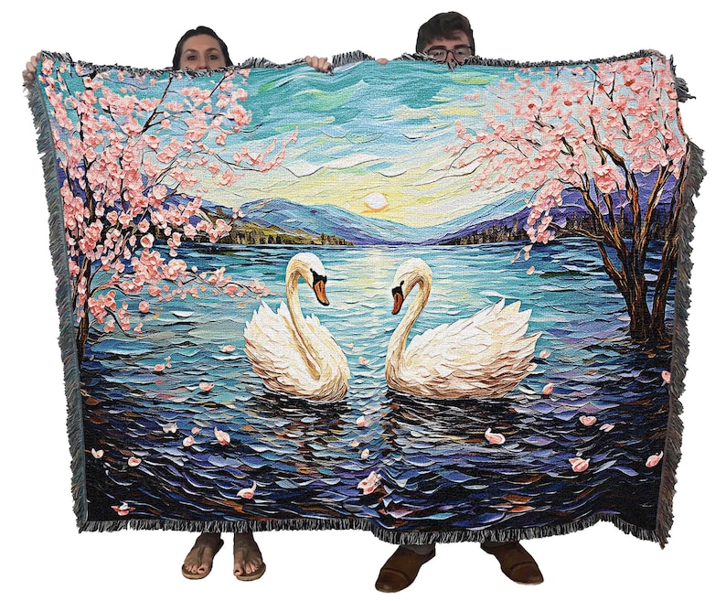 May include: A woven tapestry throw blanket features a painting of two white swans swimming in a lake with a backdrop of a blue sky, pink cherry blossoms, and a mountain range.