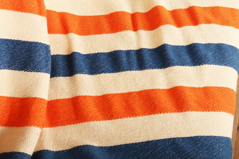 May include: A close-up of a woven textile with horizontal stripes in alternating colors. The stripes are in shades of orange, navy blue, and off-white. The fabric appears to be soft and textured, with visible weave details.
