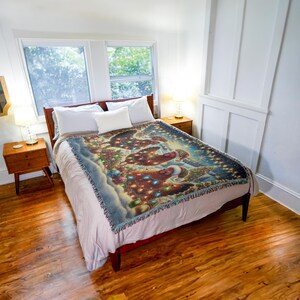 May include: A woven throw blanket featuring a festive holiday scene with three chickens wearing Santa hats, surrounded by Christmas lights, ornaments, and pine branches, draped over a bed with white pillows and a solid light-colored bedspread.