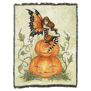 May include: A woven throw blanket featuring a whimsical Halloween design. A fairy with orange and black butterfly wings sits atop two carved jack-o'-lanterns. The fairy wears a striped dress and stockings. Green vines and leaves surround the pumpkins.