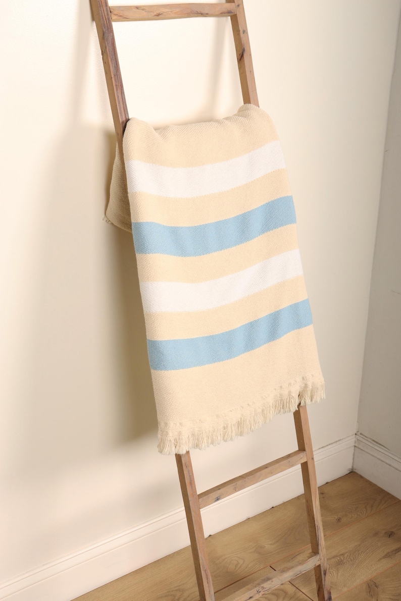 May include: A cream-colored blanket with horizontal stripes in white and light blue is draped over a wooden ladder. The blanket has a fringed edge. The ladder is leaning against a light-colored wall.