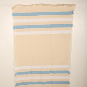 May include: A rectangular, cream-colored beach towel with horizontal stripes in light blue and white. The towel has a fringed edge along the top and bottom. The towel is hanging against a white wall.