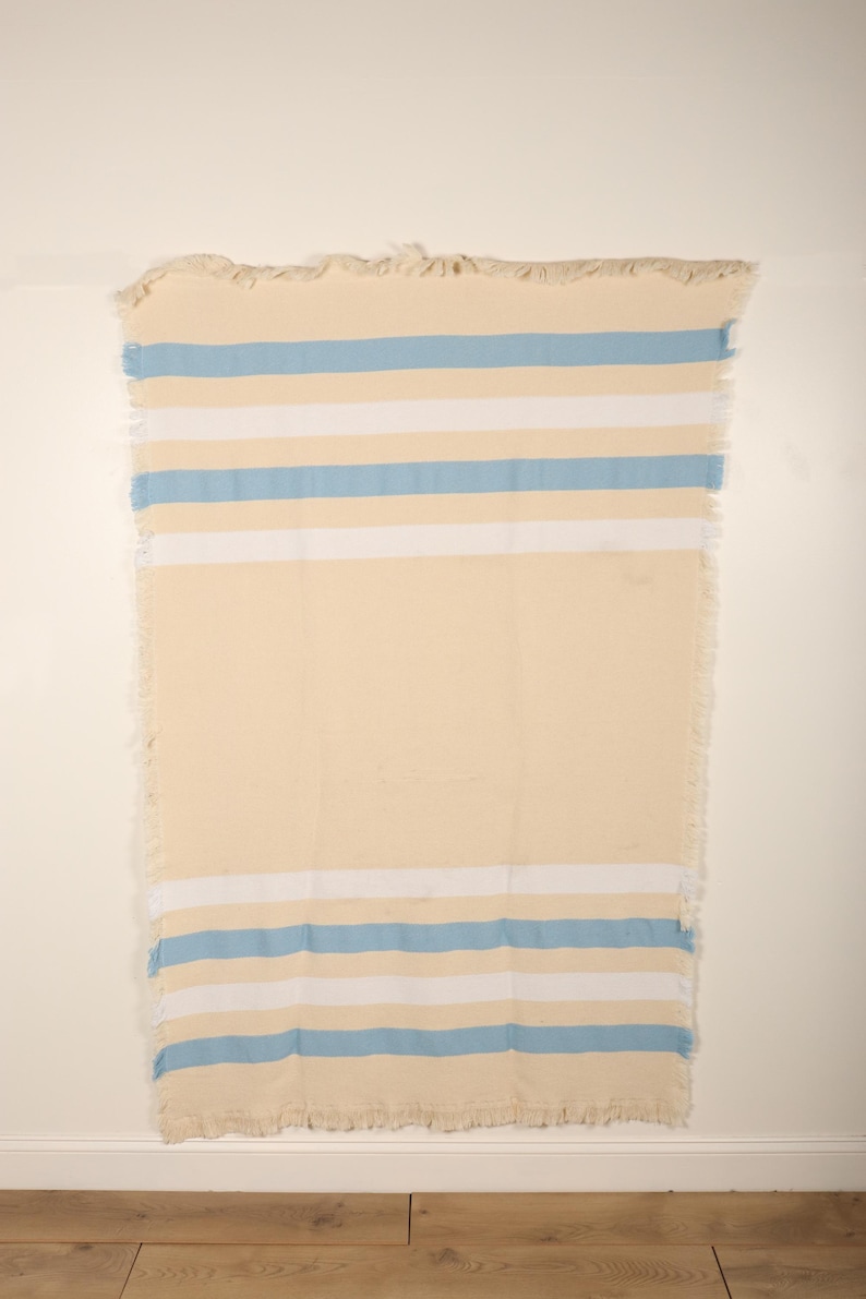 May include: A rectangular, cream-colored beach towel with horizontal stripes in light blue and white. The towel has a fringed edge along the top and bottom. The towel is hanging against a white wall.