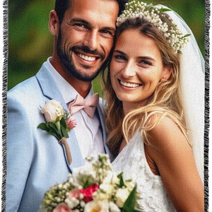 May include: A woven throw blanket featuring a smiling couple on their wedding day. The man wears a light blue suit with a pink bow tie and a rose boutonniere. The woman wears a white dress and a floral crown, holding a bouquet.