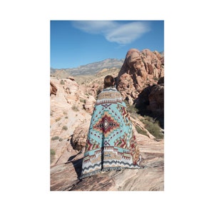 May include: A turquoise, brown, and red patterned blanket draped over a person standing on a rocky surface. The blanket features a geometric design with fringe along the bottom edge. The background shows a desert landscape with red rock formations and a blue sky.