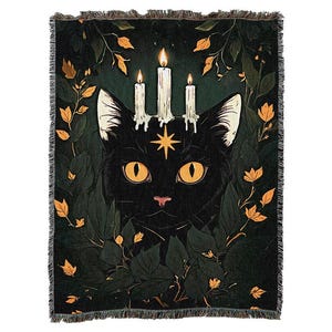Könnte beinhalten: Eine gewebte Wohndecke mit einem schwarzen Katzenmotiv, großen gelben Augen und einem goldenen Stern auf der Stirn. Zwischen den Ohren befinden sich drei brennende weiße Kerzen. Die Katze ist von dunkelgrünem Blattwerk und orangefarbenen Blättern umgeben.