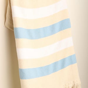 May include: A cream-colored blanket with horizontal stripes of white and light blue, draped over a wooden ladder. The blanket has a fringed edge, adding a touch of texture and detail to the soft, woven fabric.