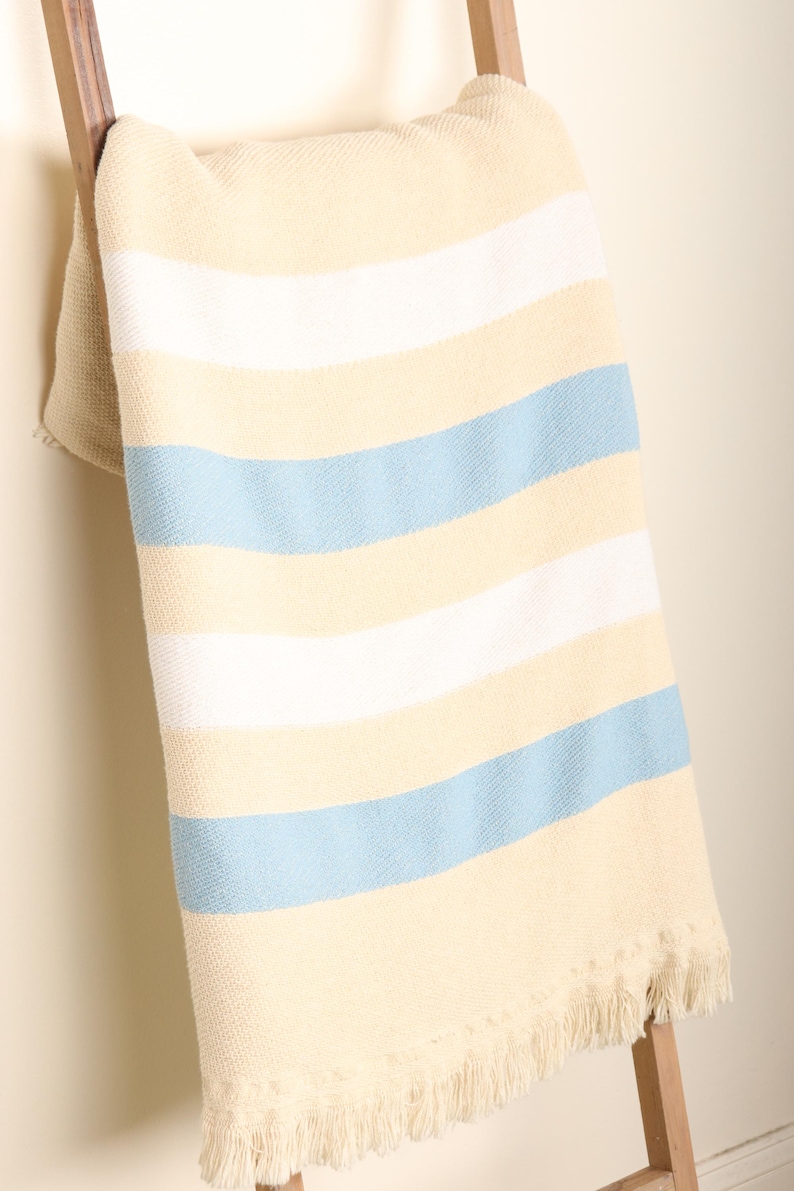 May include: A cream-colored blanket with horizontal stripes of white and light blue, draped over a wooden ladder. The blanket has a fringed edge, adding a touch of texture and detail to the soft, woven fabric.