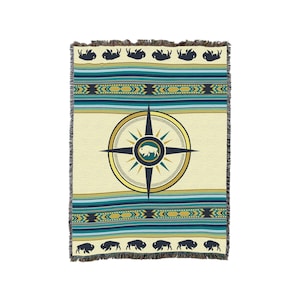 May include: A woven throw blanket with a cream background and a compass design in the center. The compass has a gold outline and a white buffalo silhouette inside. The blanket has a blue and gold geometric pattern around the compass and a fringe border with black buffalo silhouettes.