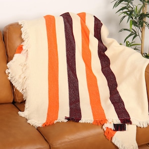 May include: A cream-colored throw blanket with vertical stripes in orange and burgundy, draped over a brown leather sofa. The blanket has fringed edges, adding a textured detail to the cozy home decor item.