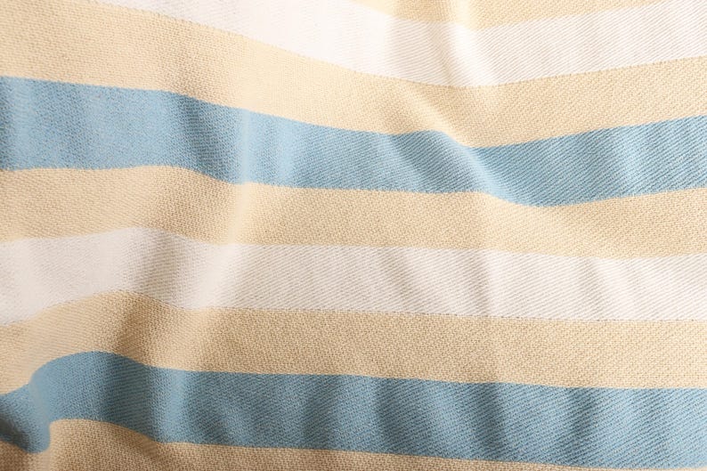May include: Close-up of a fabric with horizontal stripes in light blue, white, and tan. The fabric has a textured appearance, with the stripes running across the frame. The material appears to be soft and slightly wrinkled.