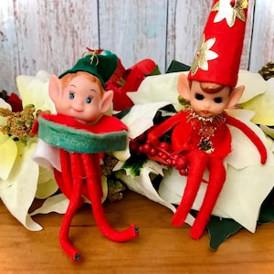 May include: Two vintage Christmas elves with red and green felt clothing. The elf on the left is holding a green and white watermelon. The elf on the right is sitting on a white poinsettia.