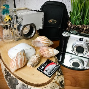 May include: A silver digital camera with a black lens and a 4GB memory card. The camera is on a silver stand and there is a clear waterproof camera case, a black camera bag, and several seashells on a wooden surface.