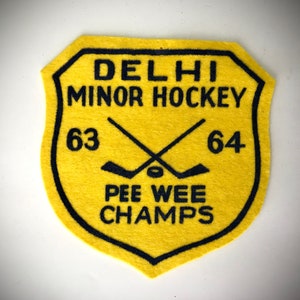 May include: Yellow felt patch with black stitching. It reads "DELHI MINOR HOCKEY 63 64 PEE WEE CHAMPS" with two crossed hockey sticks and a puck.