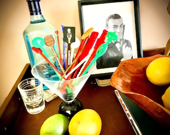 May include: A glass martini glass filled with colourful plastic cocktail stirrers. A bottle of gin, a shot glass, and a wooden bowl with lemons are also on the table.
