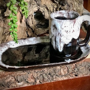 Peut inclure: Une tasse en céramique noire et blanche avec un glaçage blanc et une poignée noire repose sur un plateau ovale en céramique noire et blanche assorti. Le plateau et la tasse sont sur une surface en bois.