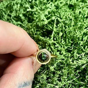 Könnte beinhalten: Ein goldener Ring mit einer kleinen, runden, transparenten Fassung. In der Fassung befindet sich ein dunkelgrüner und brauner Stein. Der Ring wird vor einem Hintergrund aus grünem Moos gehalten.