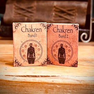 Könnte beinhalten: Zwei kleine, braune, rechteckige Schachteln mit einem dekorativen Rand. Die Schachteln sind mit "Chakren Band 1" und "Chakren Band 2" beschriftet. Jede Schachtel zeigt eine schwarze Silhouette einer Person mit einem Kreis von Symbolen um sie herum.