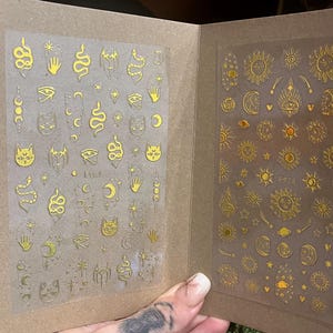 Könnte beinhalten: Goldfarbene Nagelkunst-Aufkleber mit himmlischen und mystischen Designs. Die Aufkleber zeigen Sonnen, Monde, Sterne, Schlangen, Katzen und Augensymbole. Präsentiert in einem braunen Kartonbuch.