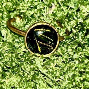 Könnte beinhalten: Ein goldener Ring mit einem runden, schwarzen Zargen, der mit einem grünen und goldenen Harzdesign besetzt ist. Das Harzdesign hat ein wirbelndes, abstraktes Muster.