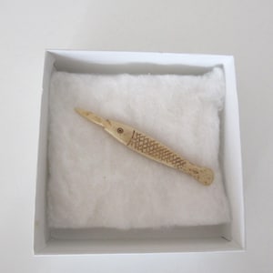 May include: A carved wooden fish-shaped object, presented in a white box with a soft, white interior. The fish has a pointed head, a textured body, a small round eye and a detailed scale pattern. The object is made of wood.