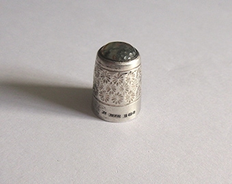 May include: A silver thimble with a floral pattern and a green stone top. The thimble is marked with the letters "M" and "B" and the number "100".