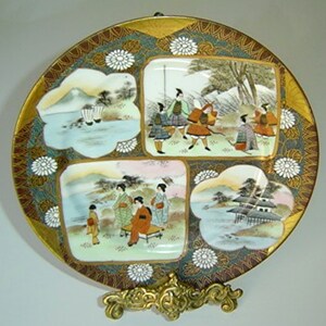 May include: A round porcelain plate with a gold rim and intricate floral designs. The plate is divided into four sections, each depicting a different scene of Japanese life. The scenes are painted in vibrant colours and feature figures in traditional clothing.