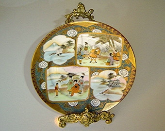 May include: A round porcelain plate with a gold rim and a blue and green floral design. The plate is divided into four sections, each featuring a different scene of Japanese people in traditional clothing. The plate is on a gold stand.