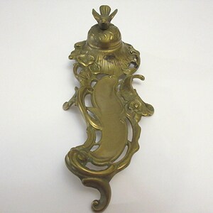 May include: An ornate brass inkwell with a bird finial and a decorative base. The inkwell features a detailed design with floral motifs and a scroll-like shape.