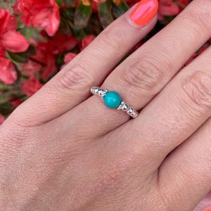 Könnte beinhalten: Ein silberner Ring mit einem türkisfarbenen Mittelstein und Perlenverzierungen. Der Ring wird vor einem Hintergrund aus roten Blumen und einer Hand mit orangefarbenem Nagellack präsentiert.