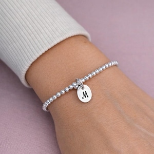 Può includere: Braccialetto con perline d'argento con un ciondolo rotondo con la lettera "M". Il braccialetto è indossato su un polso, su uno sfondo rosa tenue. Le perline sono piccole e rotonde.