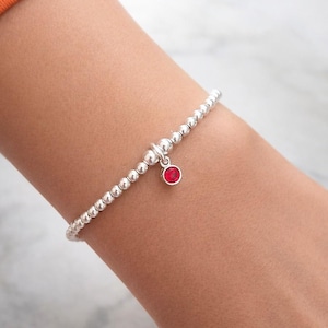 May include: A silver bracelet featuring small, round beads and a red charm. The charm is a small, round, red gemstone set in a silver bezel. The bracelet is made of small, round, silver beads strung together. The bracelet is worn on a person's wrist.