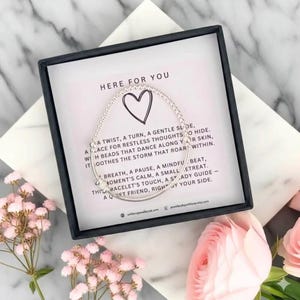 May include: A silver bracelet with a heart charm is displayed in a black box with the words "HERE FOR YOU". The box is surrounded by pink flowers. The bracelet is on a card with a poem.