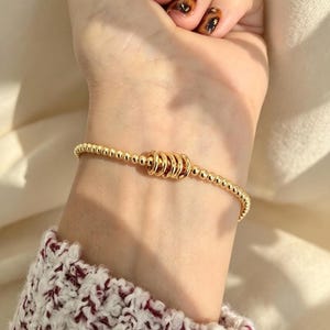 May include: A gold-toned beaded stretch bracelet featuring a series of small, uniform metallic spheres along the band, centered with a cluster of five smooth, interlocking gold-toned rings that sit horizontally across the wrist.
