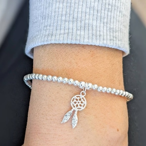 May include: A silver beaded bracelet with a silver dreamcatcher charm. The bracelet is on a person's wrist.