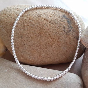 Può includere: Un braccialetto d'argento con perline, con un'unica perla più grande al centro. Il braccialetto è appoggiato su una pietra con una conchiglia accanto.