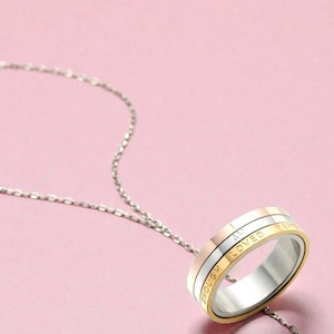 Könnte beinhalten: Eine silberne Halskette mit einem Anhängerring. Der Ring hat drei Bänder: Gold, Silber und Roségold. Die Worte "ENOUGH", "I AM LOVED" und "HAPPY" sind auf dem Ring eingraviert. Der Hintergrund ist einfarbig rosa.
