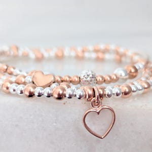 May include: A set of three bracelets with silver and rose gold beads. The bracelets are layered and feature a small heart charm.