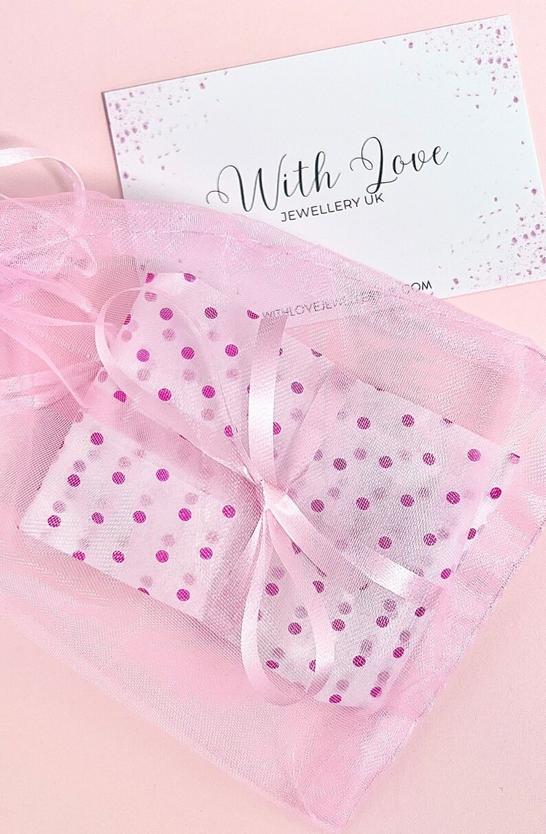 A pink polka dot gift bag with a white satin ribbon. The bag is tied with a bow and has a white card with the text 'With Love Jewellery UK' on it.