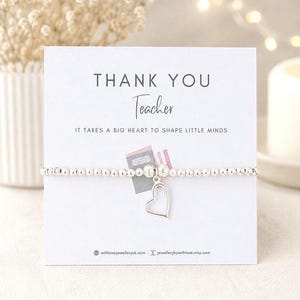 Könnte beinhalten: Ein silbernes Perlenarmband mit Herzanhänger liegt vor einer weißen Karte. Auf der Karte steht "THANK YOU Teacher" und "IT TAKES A BIG HEART TO SHAPE LITTLE MINDS". Im Hintergrund ist eine Vase mit weißen Blumen zu sehen.