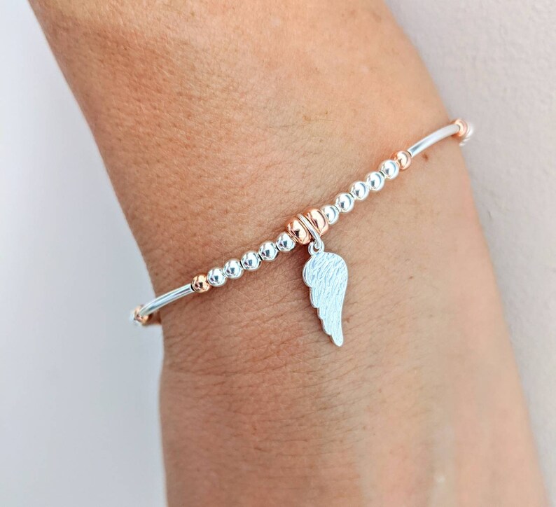 Puede incluir: Una pulsera de cuentas de plata y oro rosa con un dije de ala de &aacute;ngel de plata. La pulsera est&aacute; en la mu&ntilde;eca.