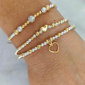 May include: Three beaded bracelets in gold and silver tones. One bracelet features a sparkling ball, another a gold heart, and the third a dangling heart charm. The bracelets are worn on a wrist.