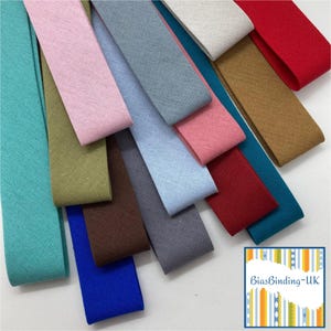 May include: A selection of colourful bias binding strips in shades of teal, pink, blue, and red. The fabric strips are arranged diagonally, displaying their different hues and textures. The image includes a logo in the bottom right corner.